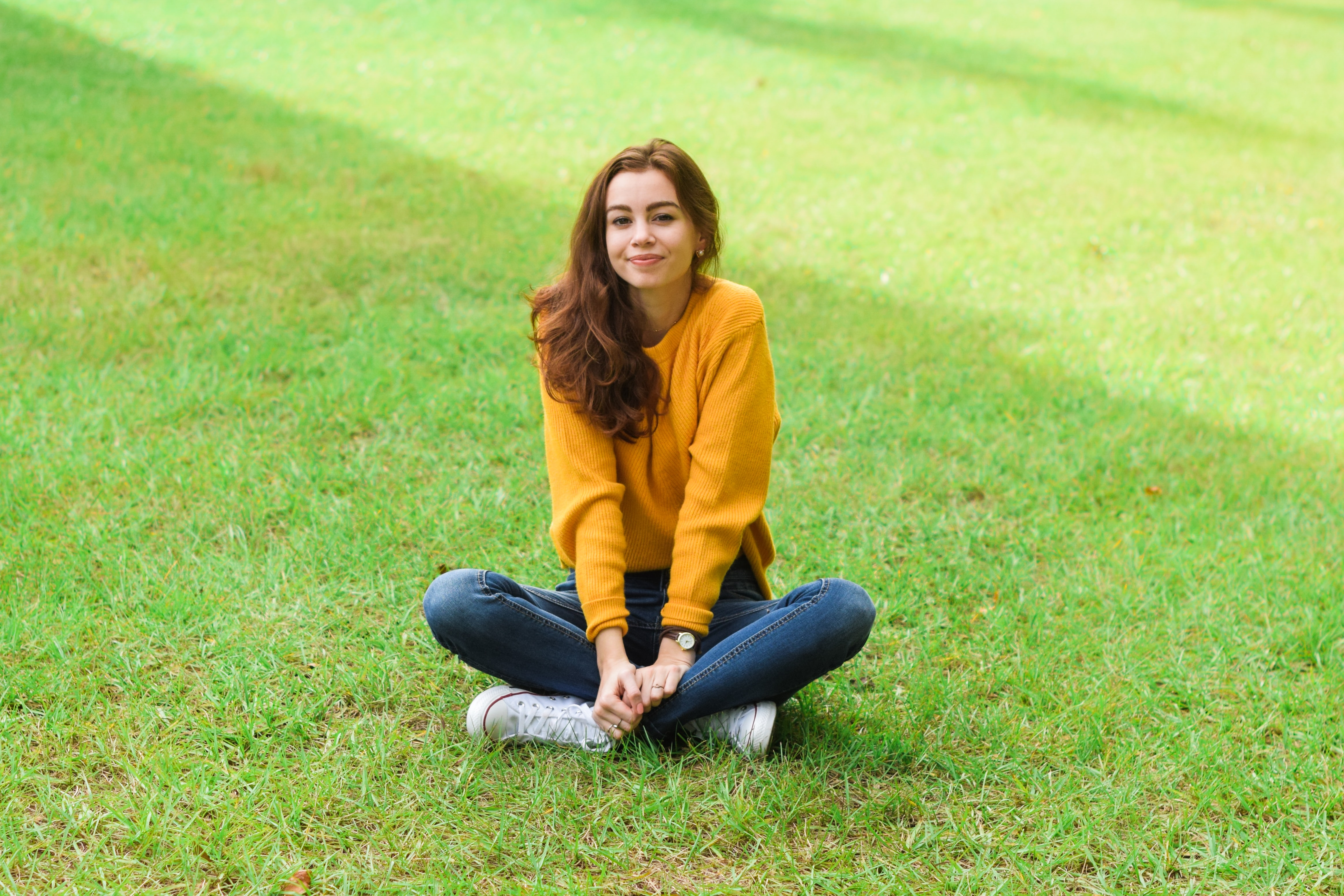 A young lady sitting cross-legged in the middle of a green lawn. She is wearing a mustard yellow sweater, blue jeans, and white sneakers.