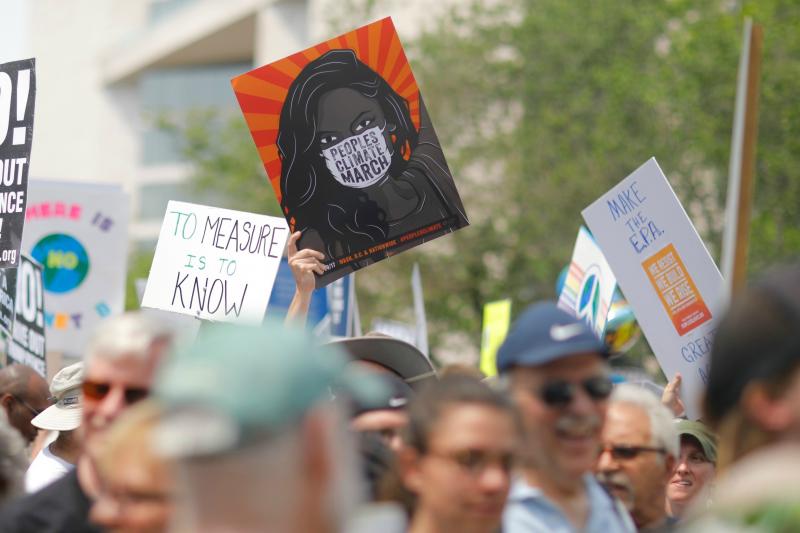 The image is of a march for climate change, many signs are raised, and people of all ages are shown. 