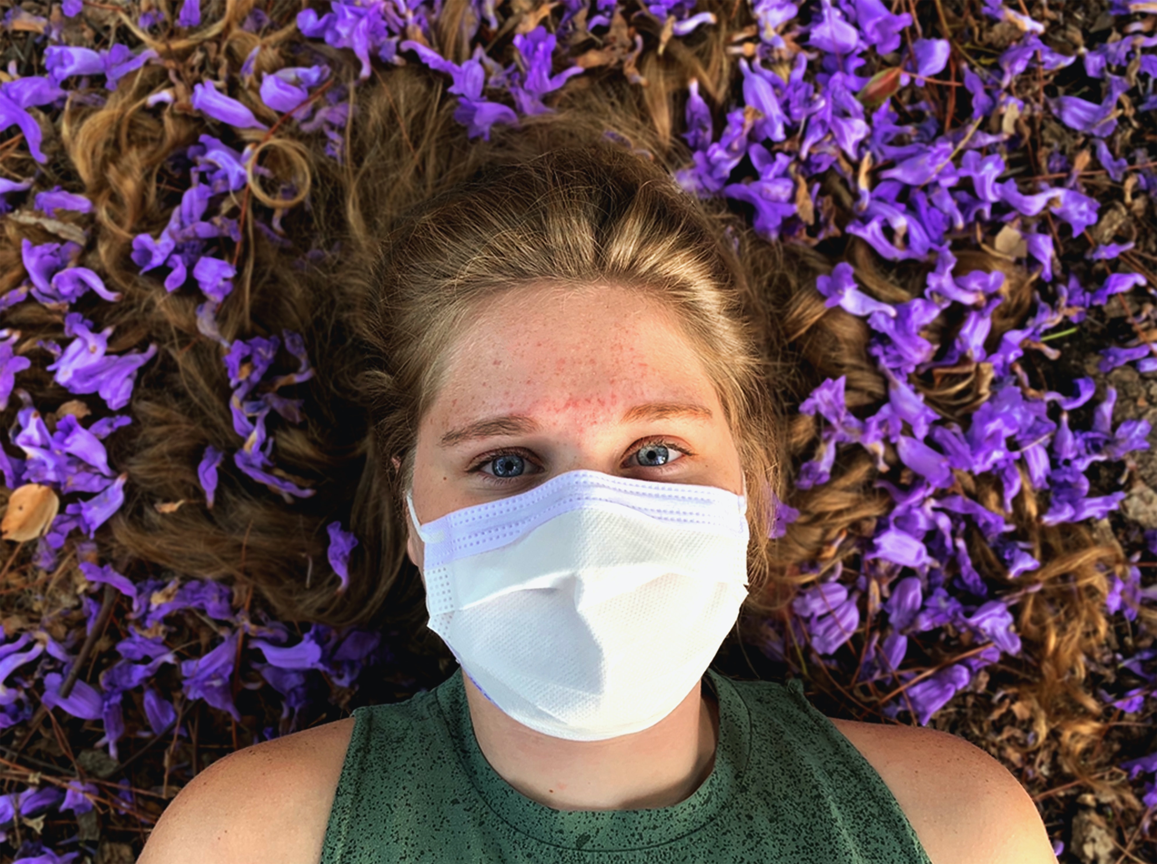 A young woman lays in a bed of purple flowers with her hair splayed out around her head. She is wearing a green, sleeveless top and a white mask. 