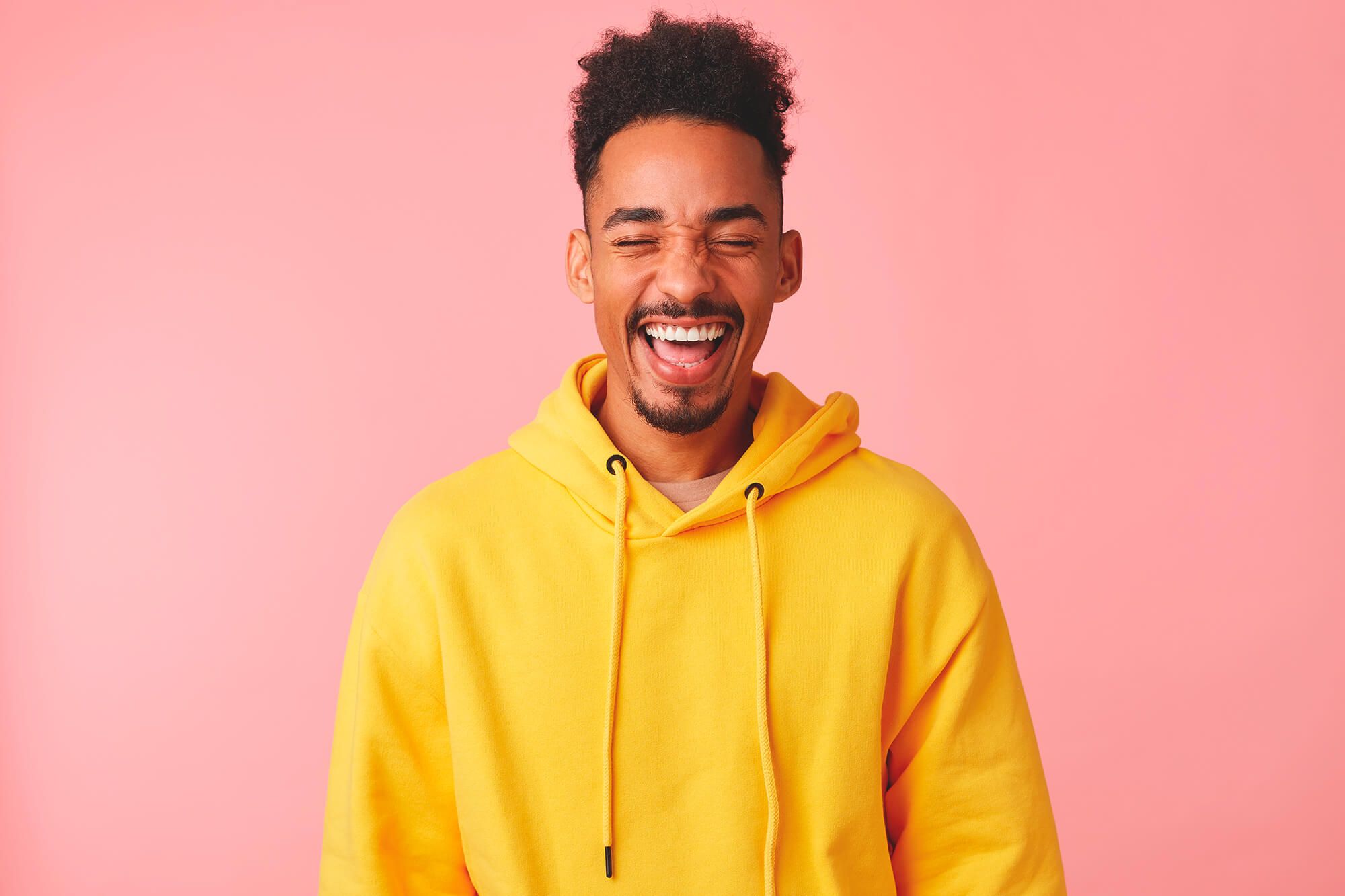 A young man, laughing, stands in front of a pink background. He's wearing a yellow sweater. 