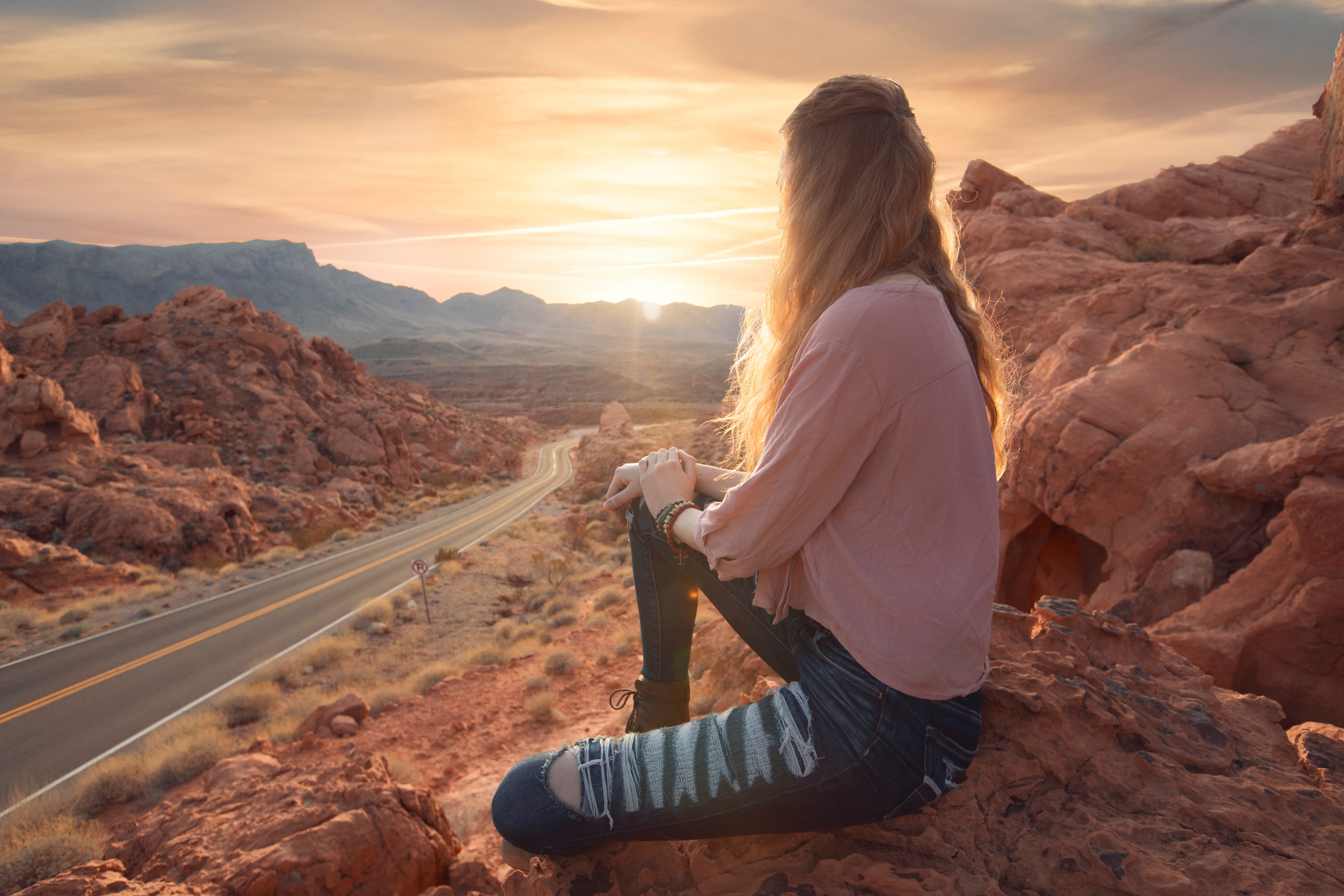 A young woman sits on a mountainside staring into the distance, where the sun is rising. 