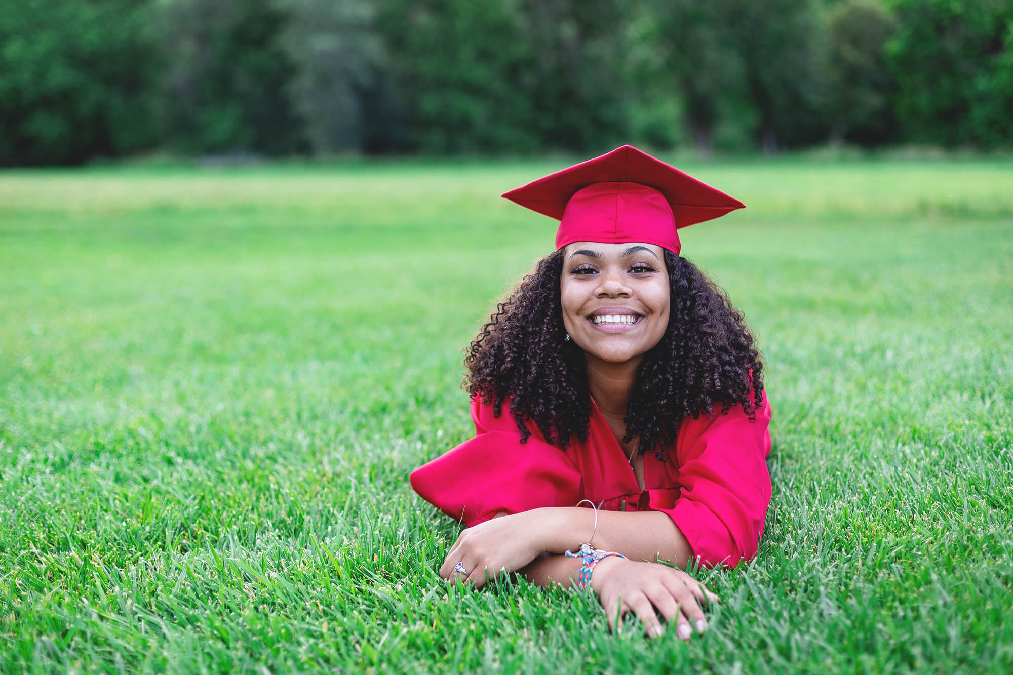 a young woman lays in the grass with her red cap and gown on