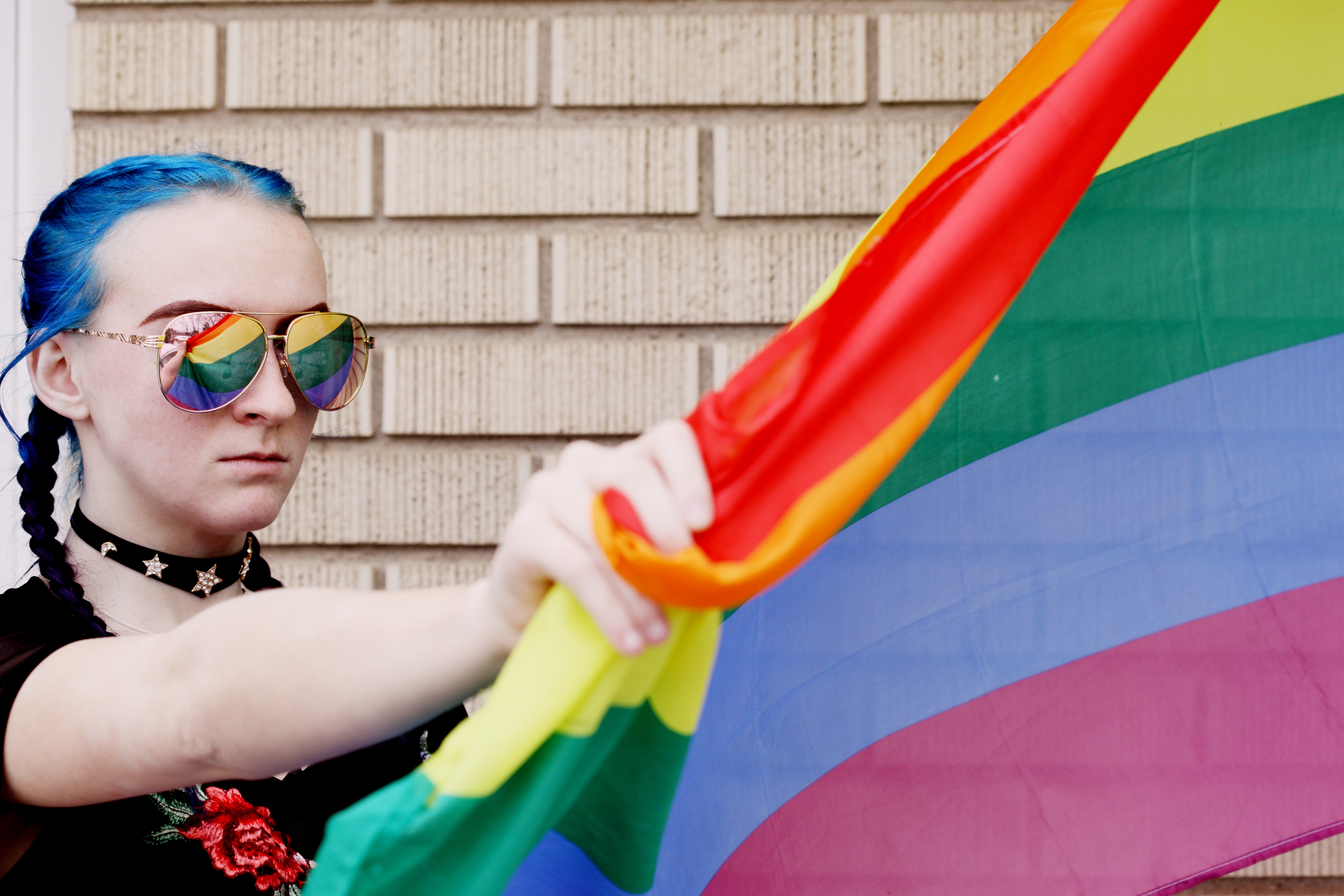 Student with blue hair in reflective silver sunglasses holds a gay pride flag.