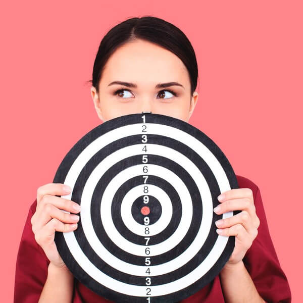 A young woman holds a dart board. She is in front of a fun pink background. 