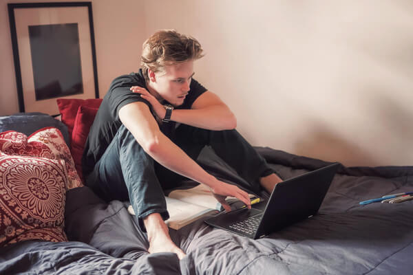 a man sits on his bed looking at his laptop