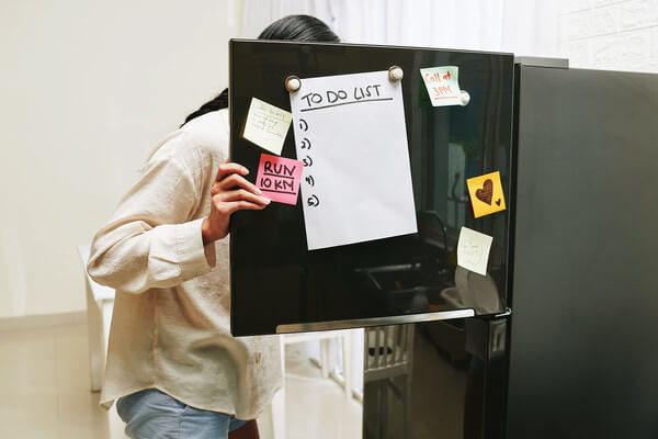a student looks inside of a refrigerator with a to do list hanging on the door. 