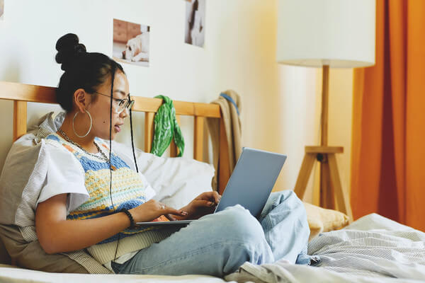 a young woman sits on her bed looking at colleges on her laptop