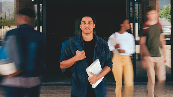 a student in a blue button down shirt with a backpack over one should and a white book in the other hand standing in front of a building