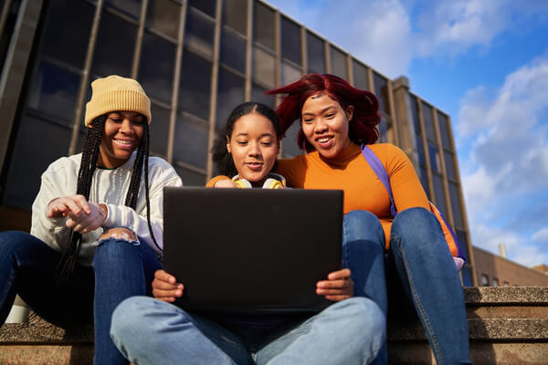 3 female students sitting outside on the steps looking at one black laptop
