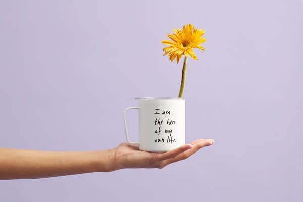 A person holding a mug with a yellow flower in it that says, "I am the hero of my own life."